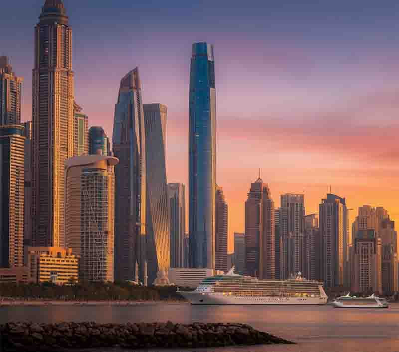 Skyline bei Sonnenuntergang in Dubai. Im Vordergrund liegt ein großes, weißes Kreuzfahrtschiff auf dem Wasser, mit einem kleineren Schiff rechts davon. Das Ciel Dubai Marina Hotel dominiert den Hintergrund.