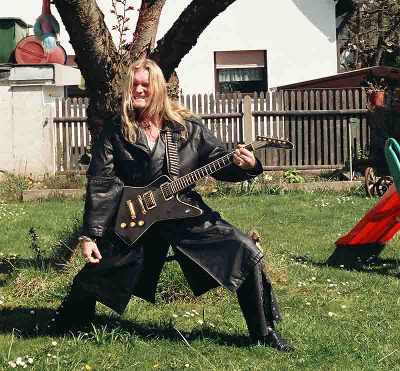 Ein Heavy Metal Fan mit langen blonden Haaren und langem Ledermantel posiert mit einer schwarzen E-Gitarre in seinem grünen Garten.